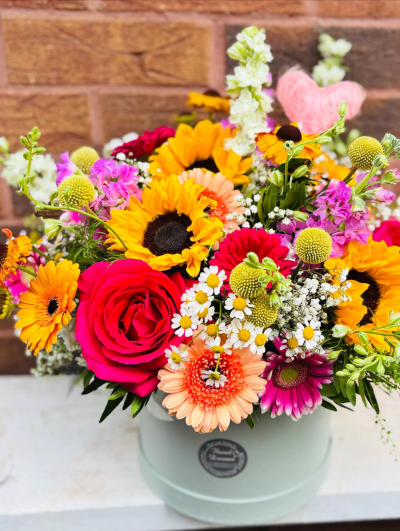Happy Hatbox - A joyful burst of colour featuring vibrant sunflowers and lively seasonal blooms. Arranged in a soft pastel hatbox, this cheerful display blends sunny yellows, fiery oranges, vivid pinks, and fresh greens.